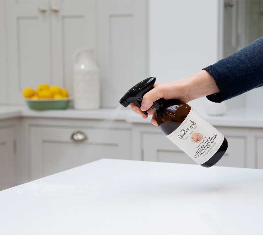 Person holding a bottle of greenscents product over a white surface in a kitchen setting.