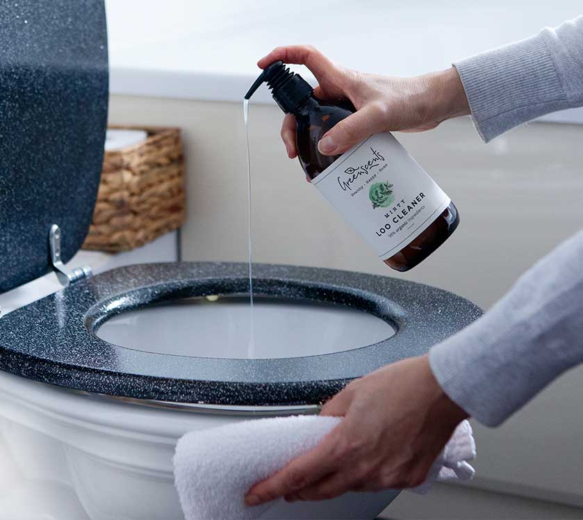 Person cleaning a toilet with a spray bottle labeled 'Greenscents' and a cloth.
