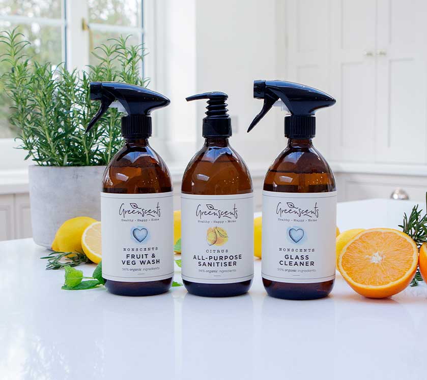 Three bottles of greenscents cleaning products on a kitchen counter with fruits and plants.