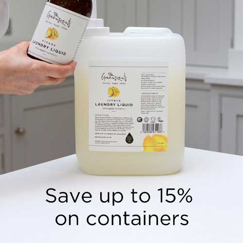 Hand holding a bottle of greenscents laundry liquid next to a large container with a label, set against a kitchen background.