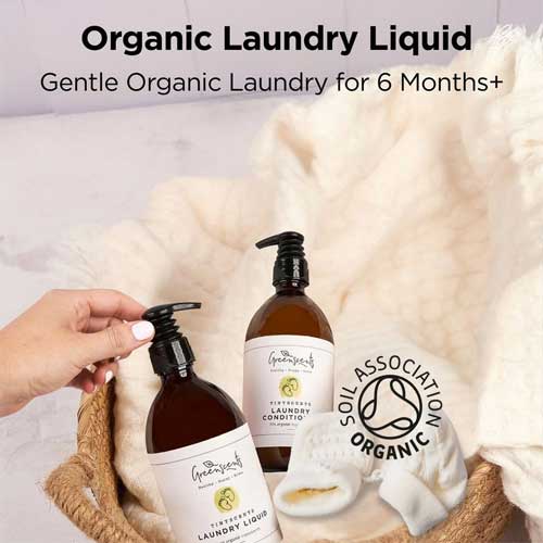 Two bottles of greenscents organic laundry liquid with a hand holding one bottle, placed on a textured surface.