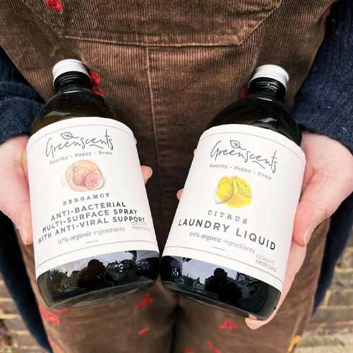 Two bottles of Greenscents products held by a person wearing brown pants.