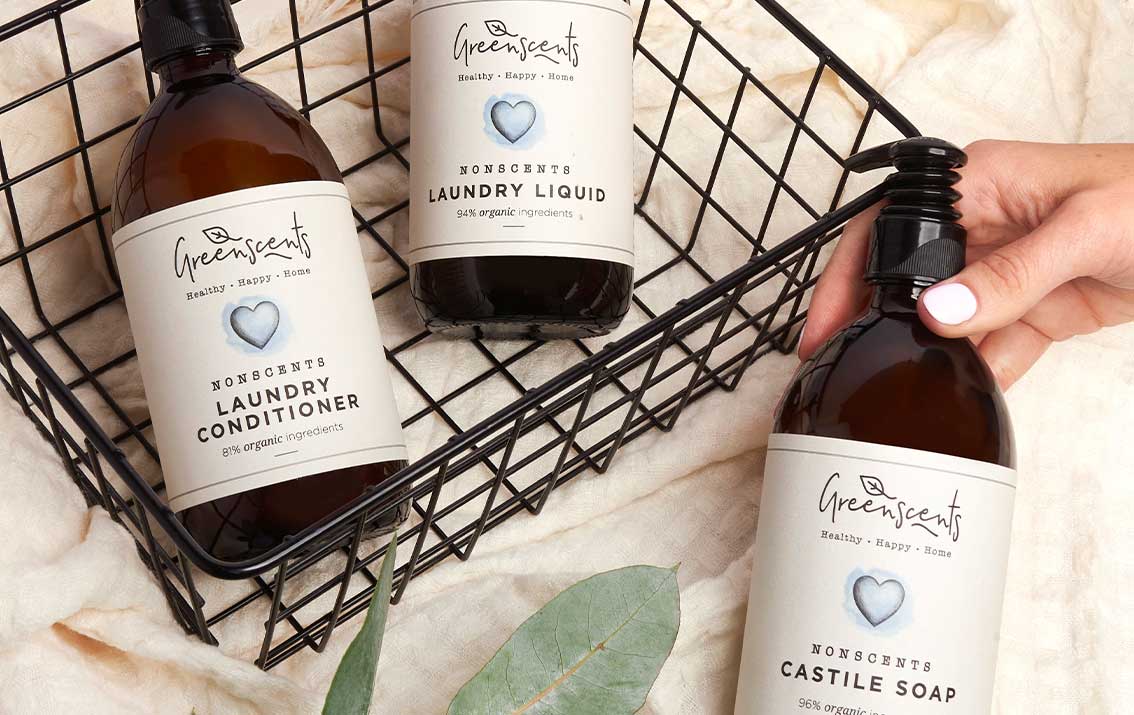 Three bottles of GreenScents laundry products on a wire basket with a hand holding one bottle.