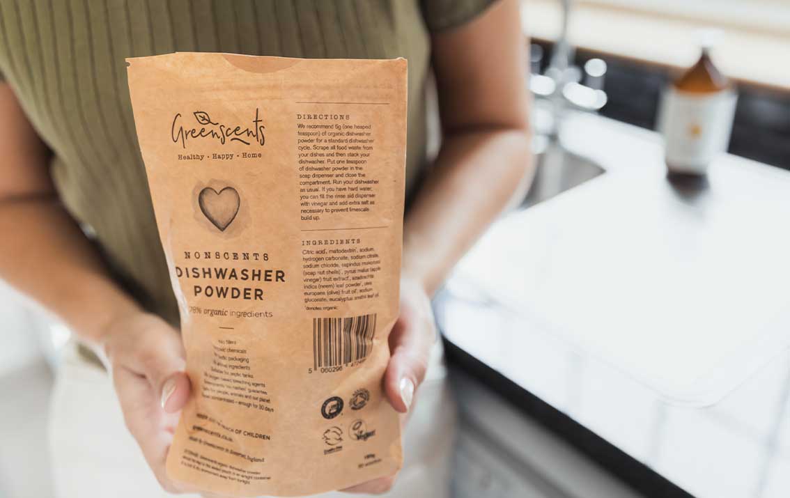 Person holding a package of Greenscents dishwasher powder in a kitchen.