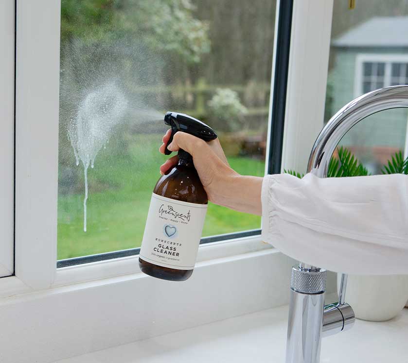 Person cleaning a window with a spray bottle labeled greenscents in a bathroom setting.
