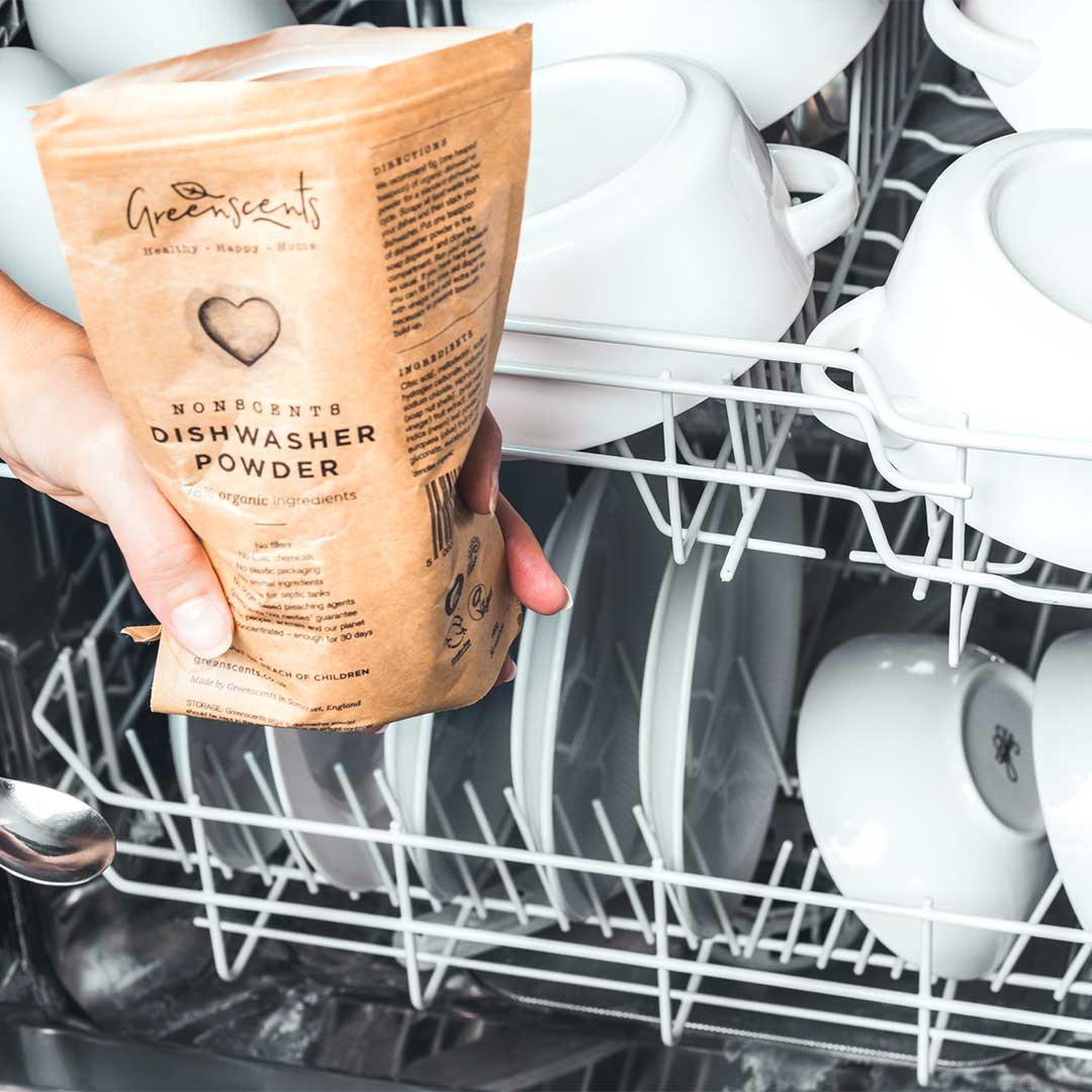 Person holding a package of Greenscents dishwasher powder in front of a dishwasher filled with clean dishes.