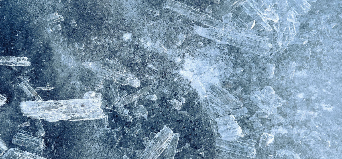 A close-up texture of frozen ice or snow with visible translucent crystal structures and fine frosty detail in cool blue and white tones.