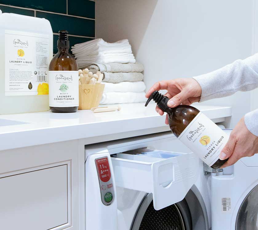 Person holding a bottle of greenscents laundry detergent above a washing machine in a laundry room.