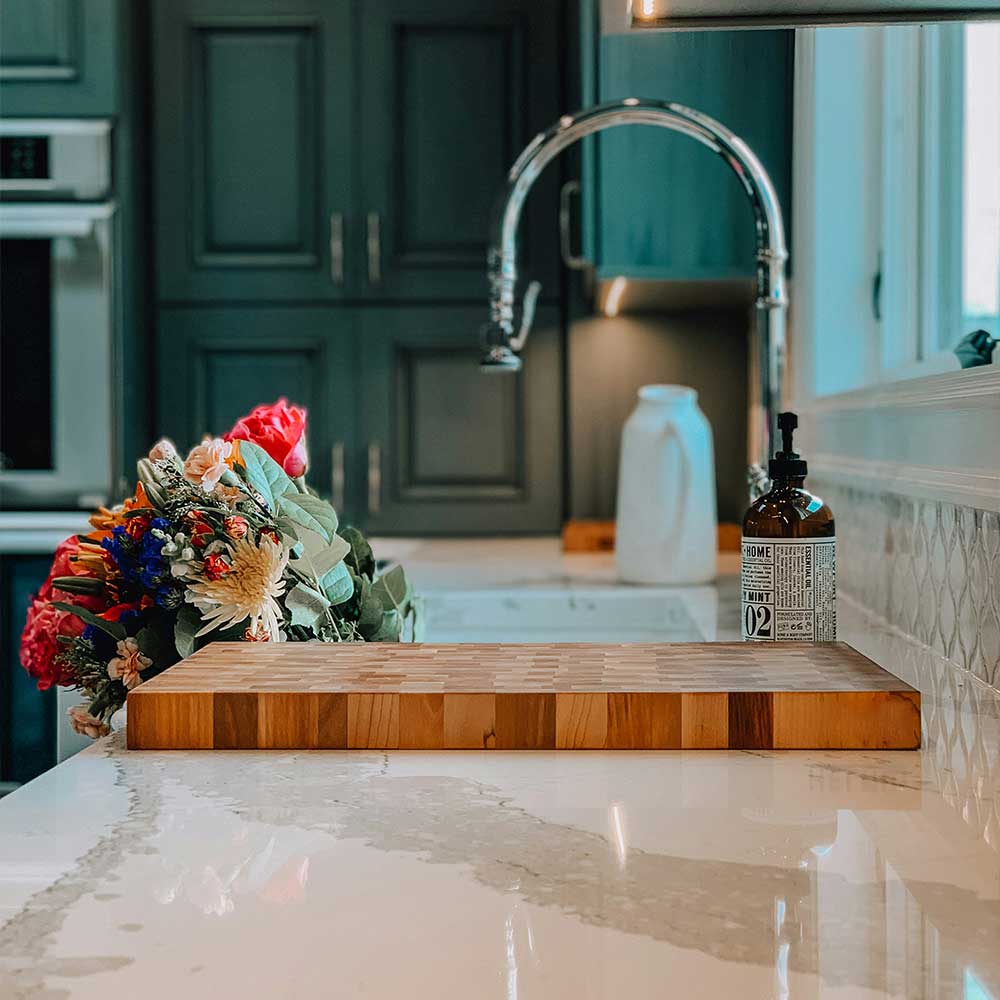 Wooden cutting board on a kitchen counter with a bottle and flowers in the background.