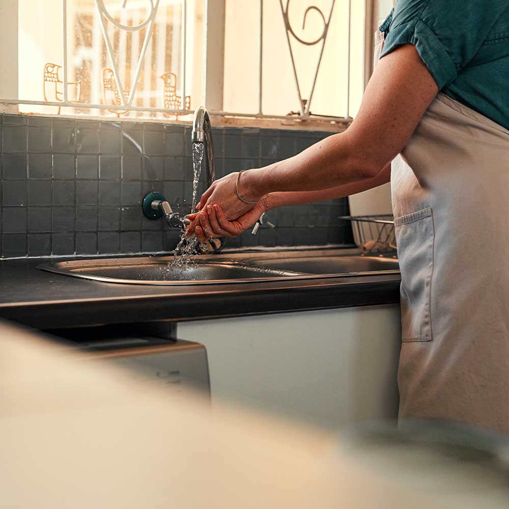Person washing hands under a running faucet in a kitchen.