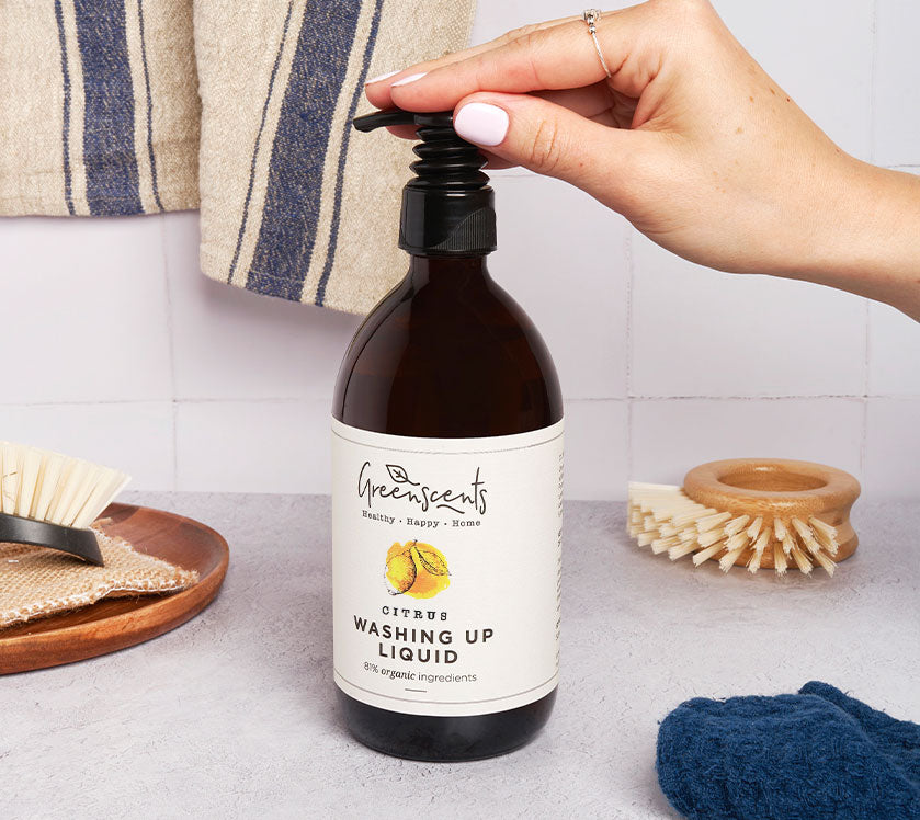 Hand holding a bottle of GreenScents washing up liquid on a kitchen counter.